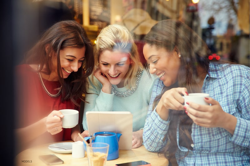 Collection of three women, smiling, drinking coffee, and looking at tablet screen in a shop.